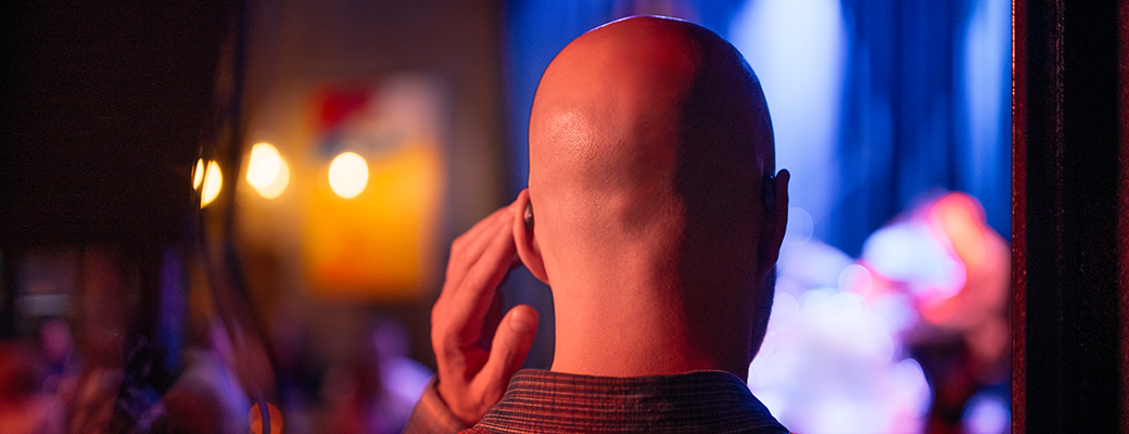 Behind view of male hearing aid wearer touching hearing aid while looking at a musician on stage